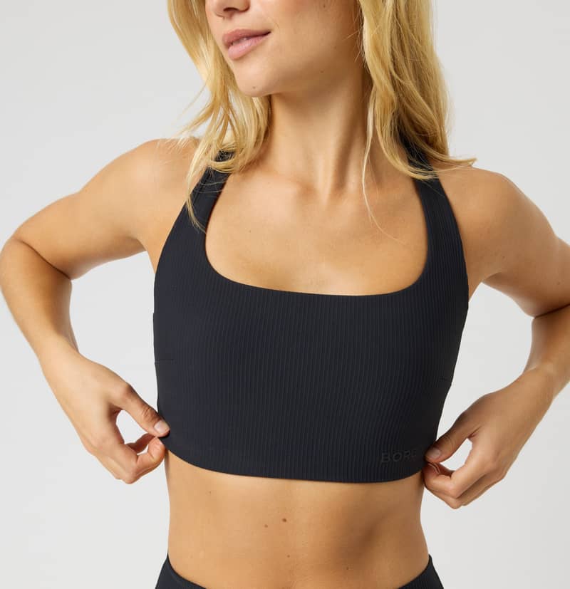 A woman with long blonde hair wearing a black sports bra and matching bottoms, adjusting the fit while posing against a plain background.