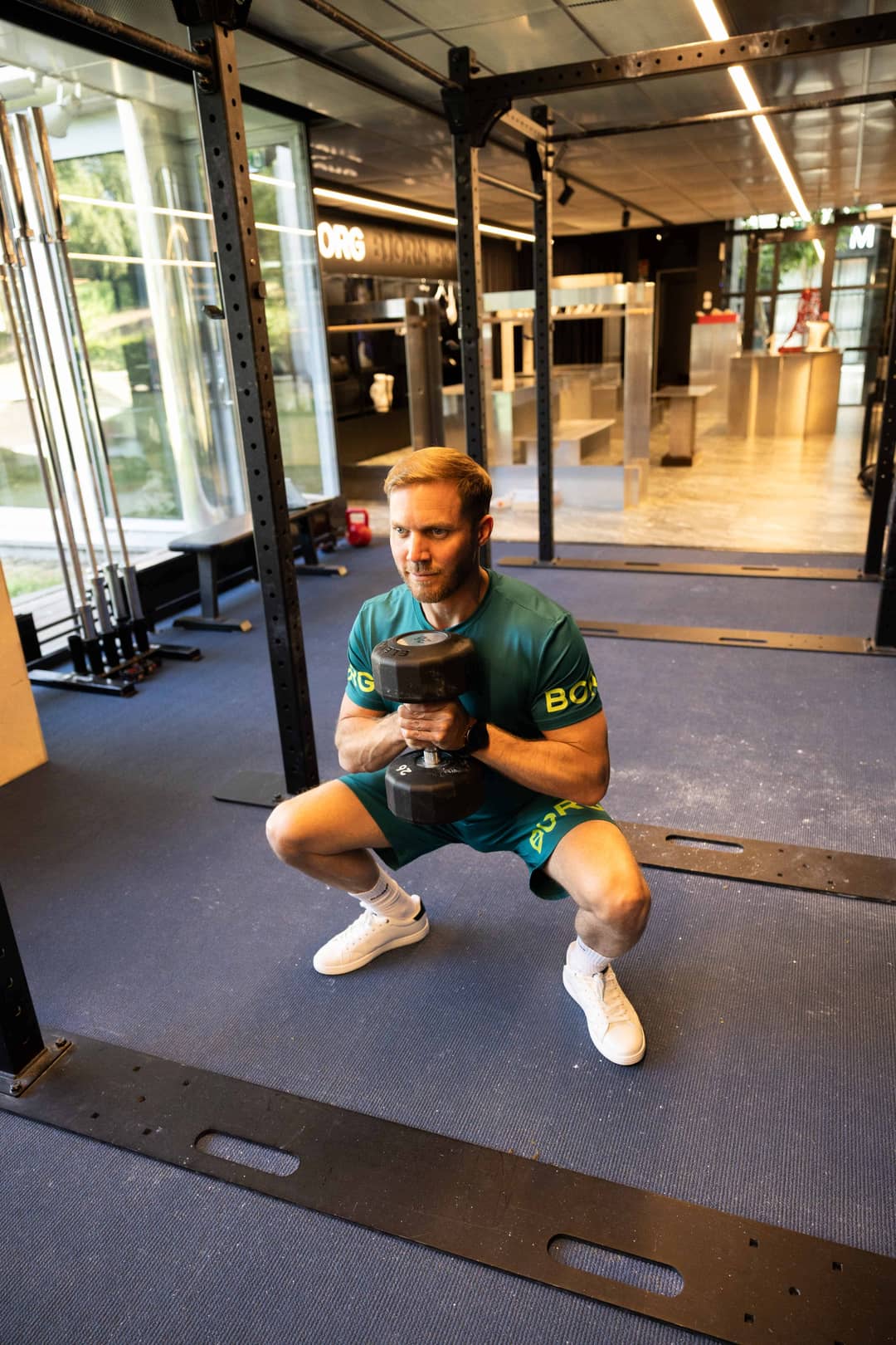 Man doing a goblet squat with a dumbbell, wearing a green workout set and white sneakers from Björn Borg