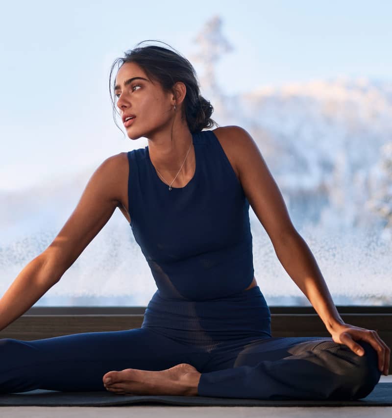 A woman in a navy workout outfit sits cross-legged indoors, with a snowy landscape visible through the window behind her.