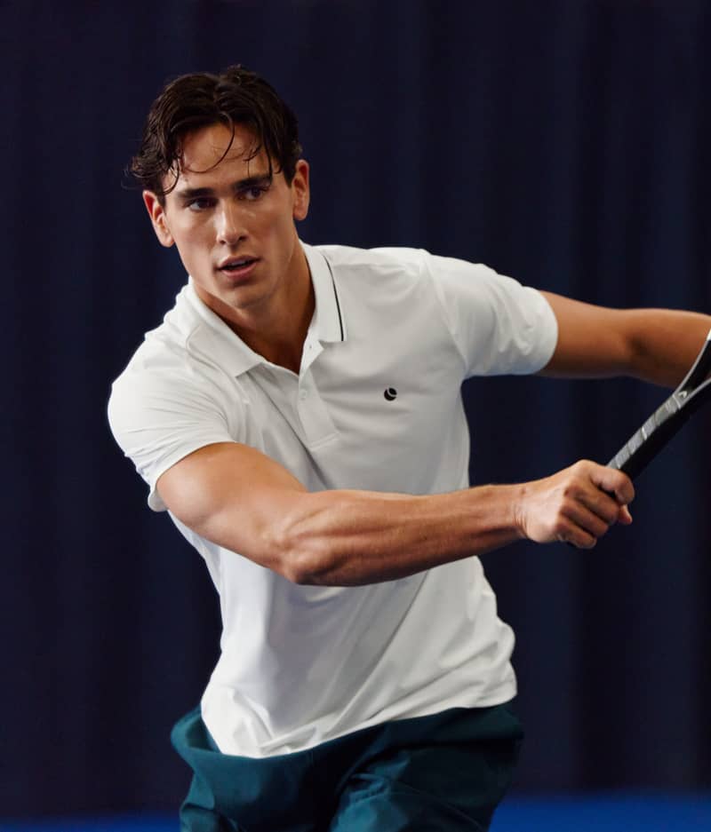 A man in a white polo shirt playing tennis indoors, holding a racket, focused on the ball.