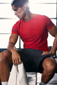 Man sitting on a workout bench wearing a red training T-shirt and dark shorts, holding a white towel.