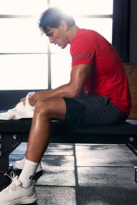 Athlete in a red shirt sits on a bench in a gym, holding a towel, with bright sunlight streaming through a window behind him.