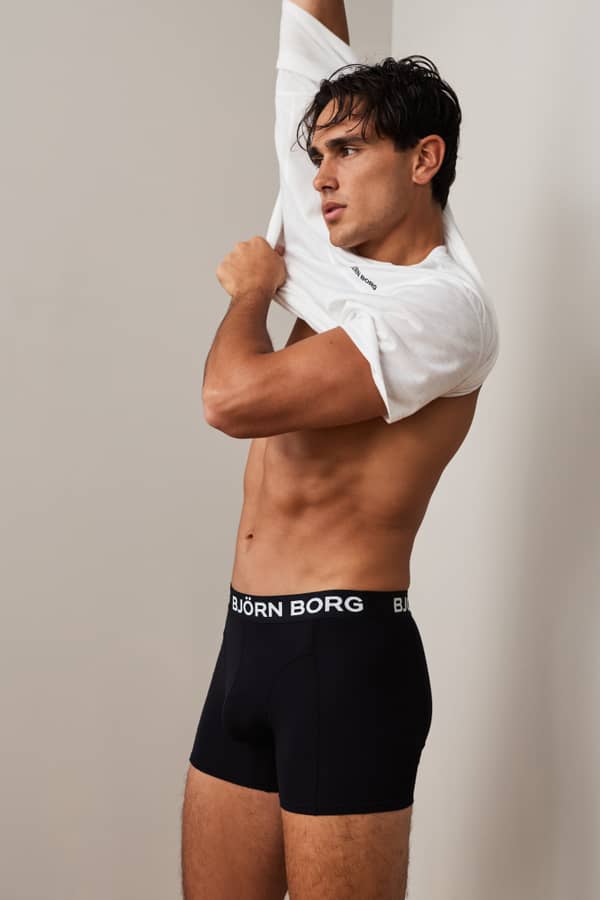 Man changing into a white T-shirt while wearing black boxer briefs indoors