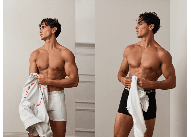 A shirtless man in white underwear on the left and black underwear on the right, holding a garment in each hand, standing indoors.