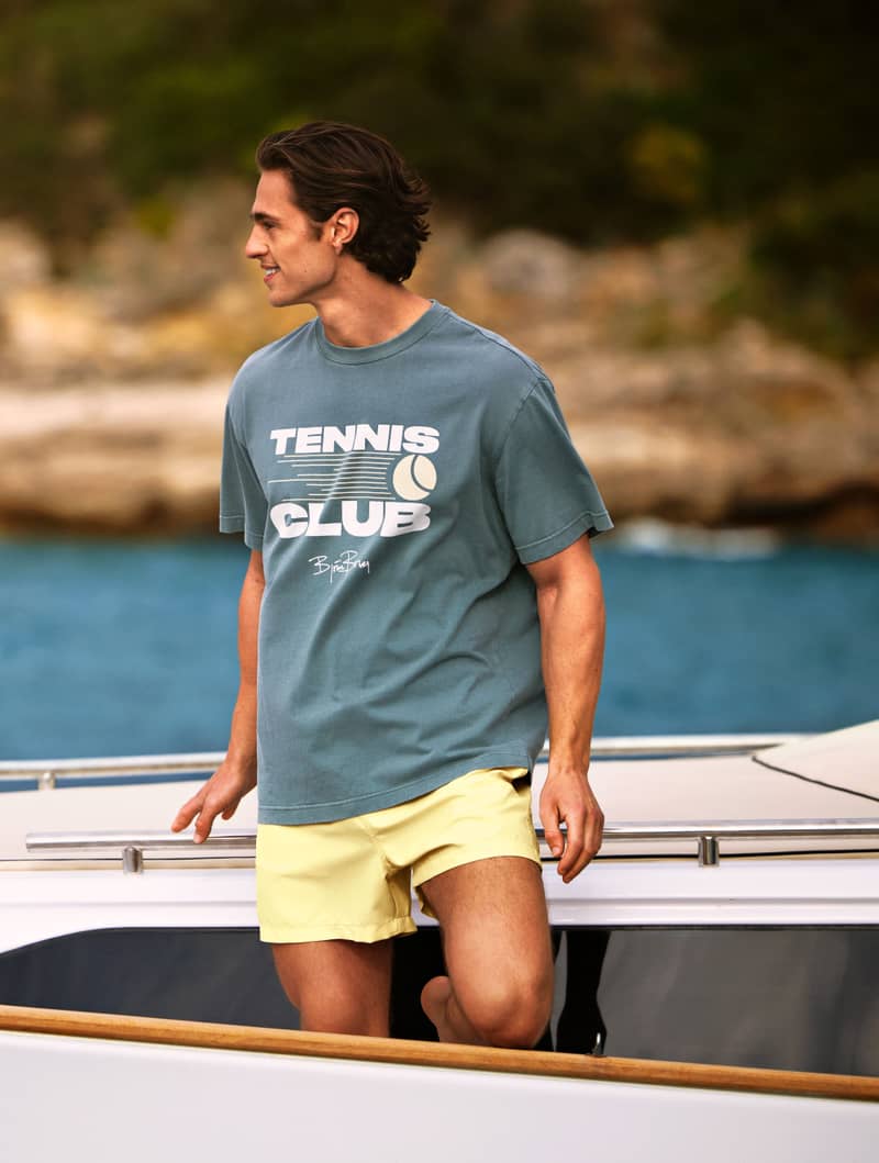A man in a "Tennis Club" shirt and yellow shorts stands on a boat, with a rocky shoreline and blue water in the background.