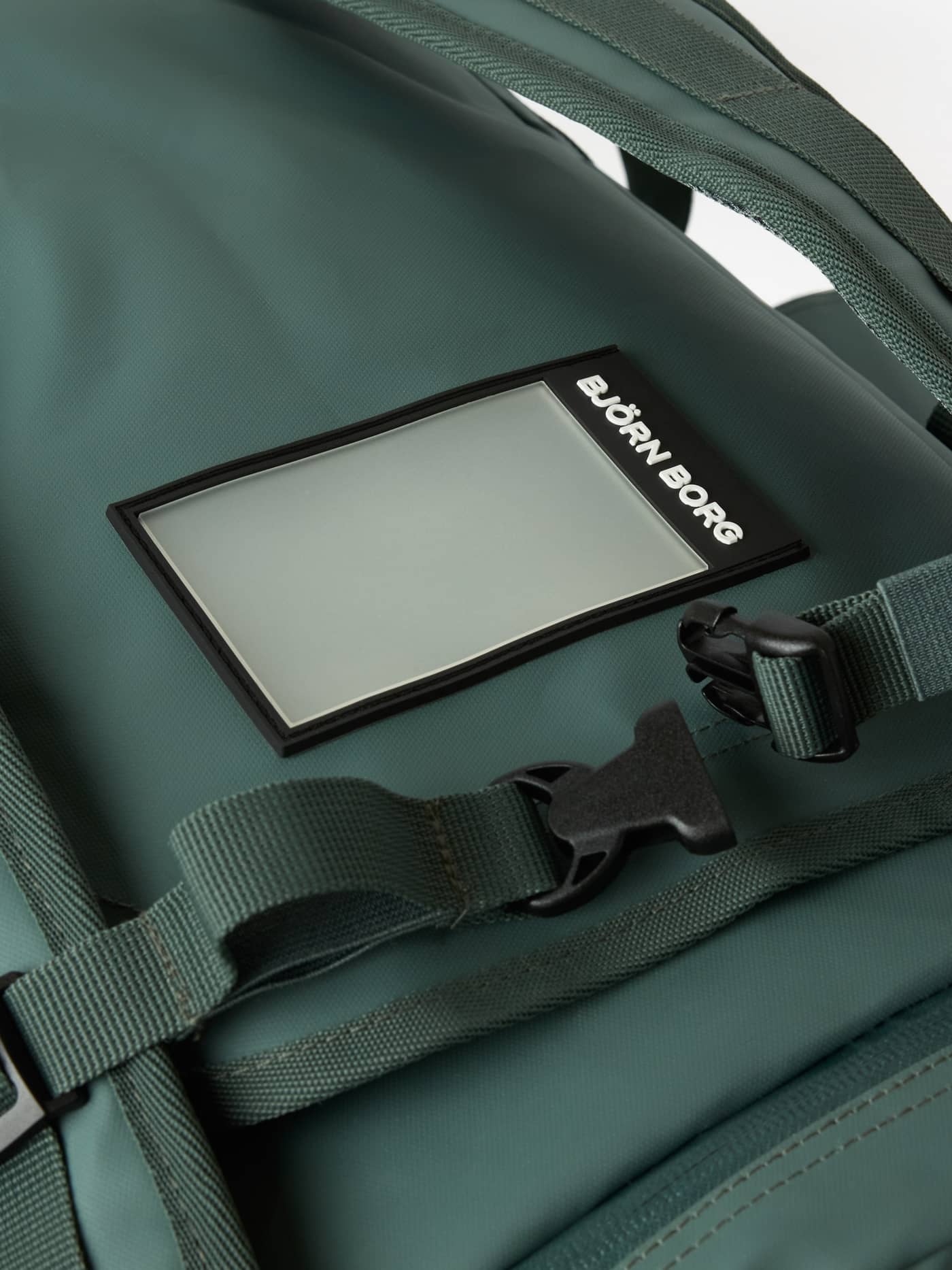 Close-up of the black address tag with white logo and buckle on the Balsam Green (Green) Borg Duffle Bag 35L. - Image 6
