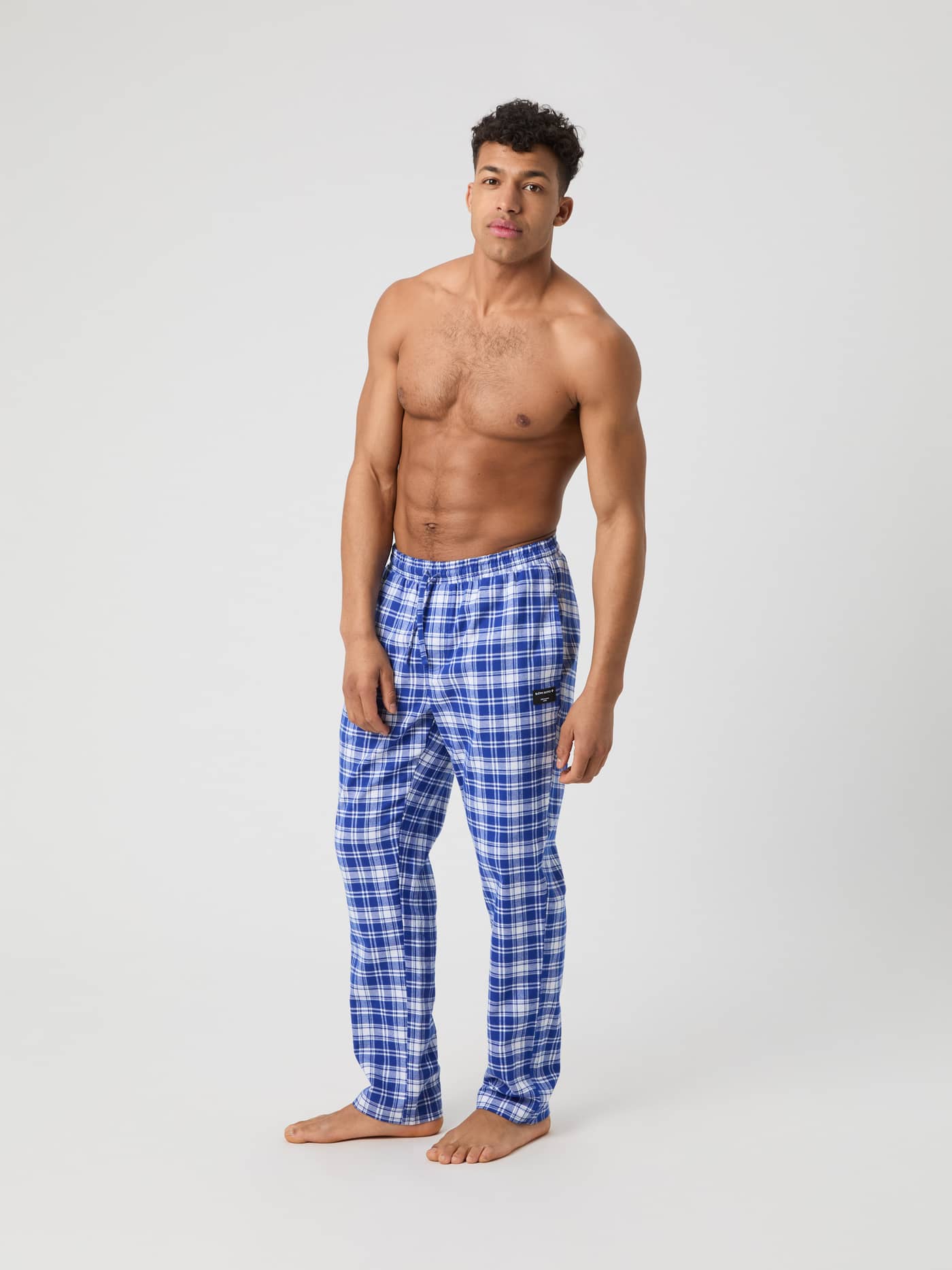 A male model stands shirtless, wearing the Björn Borg Core Flannel Pyjama Pants in a blue and white check pattern. - Image 1
