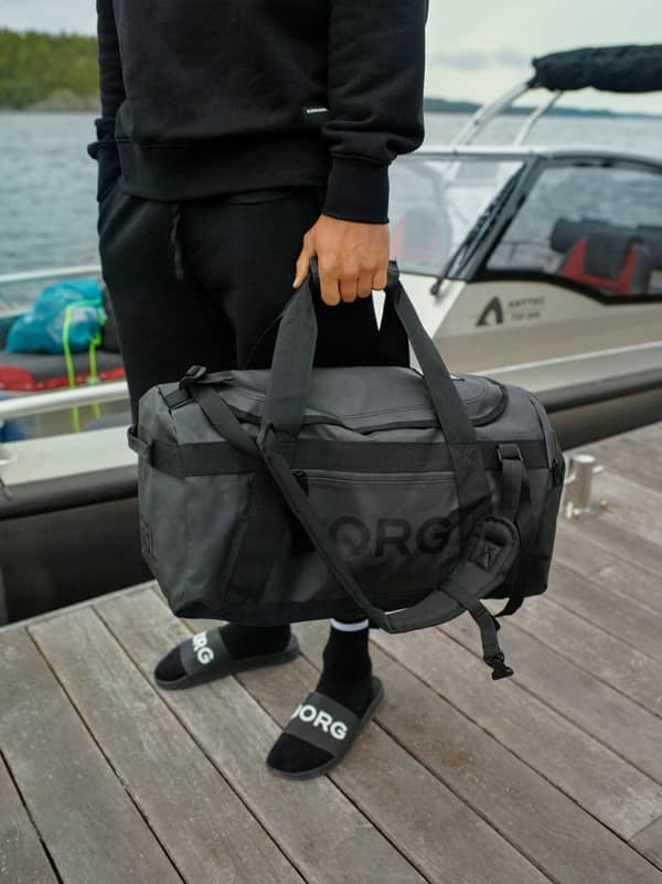 Borg Duffle Bag 55L in Black Beauty (Black) with a black logo, held by a person on a wooden dock by the water.