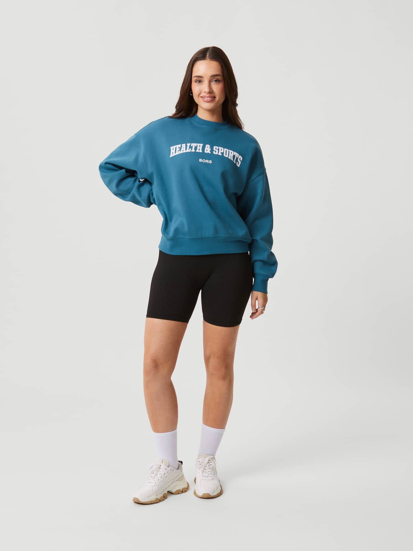 Full-body front view of a woman wearing the Studio Oversized Crew in Mallard Blue (Blue) with a white chest logo print. - Image 4