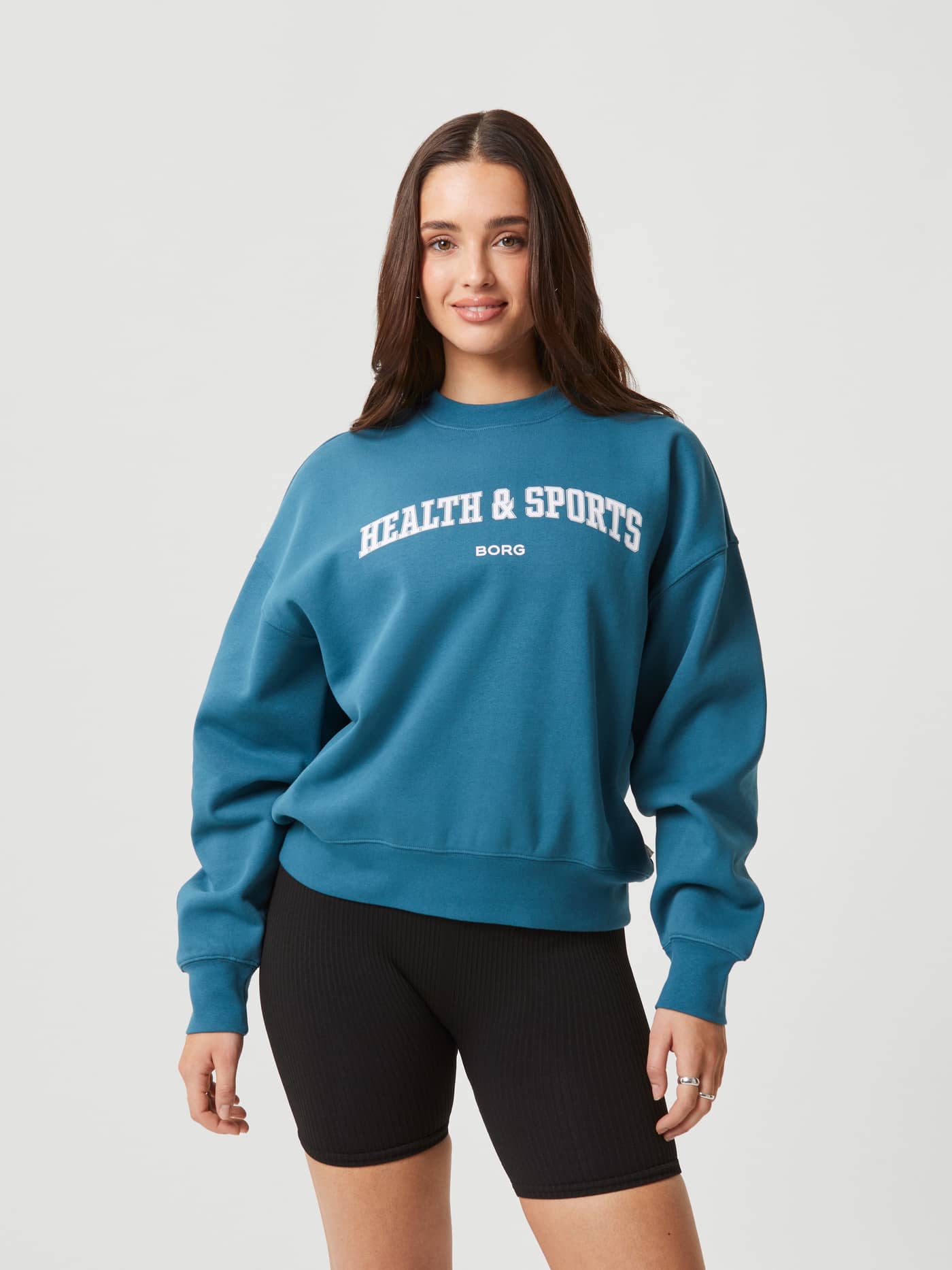 Front view of a woman in the Björn Borg Studio Oversized Crew in Mallard Blue (Blue) with an oversized fit and white logo. - Image 2