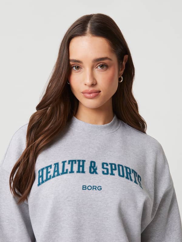 Studio Oversized Crew i Light Grey Melange (Grå) set forfra i nærbillede med et blågrønt brystlogo.