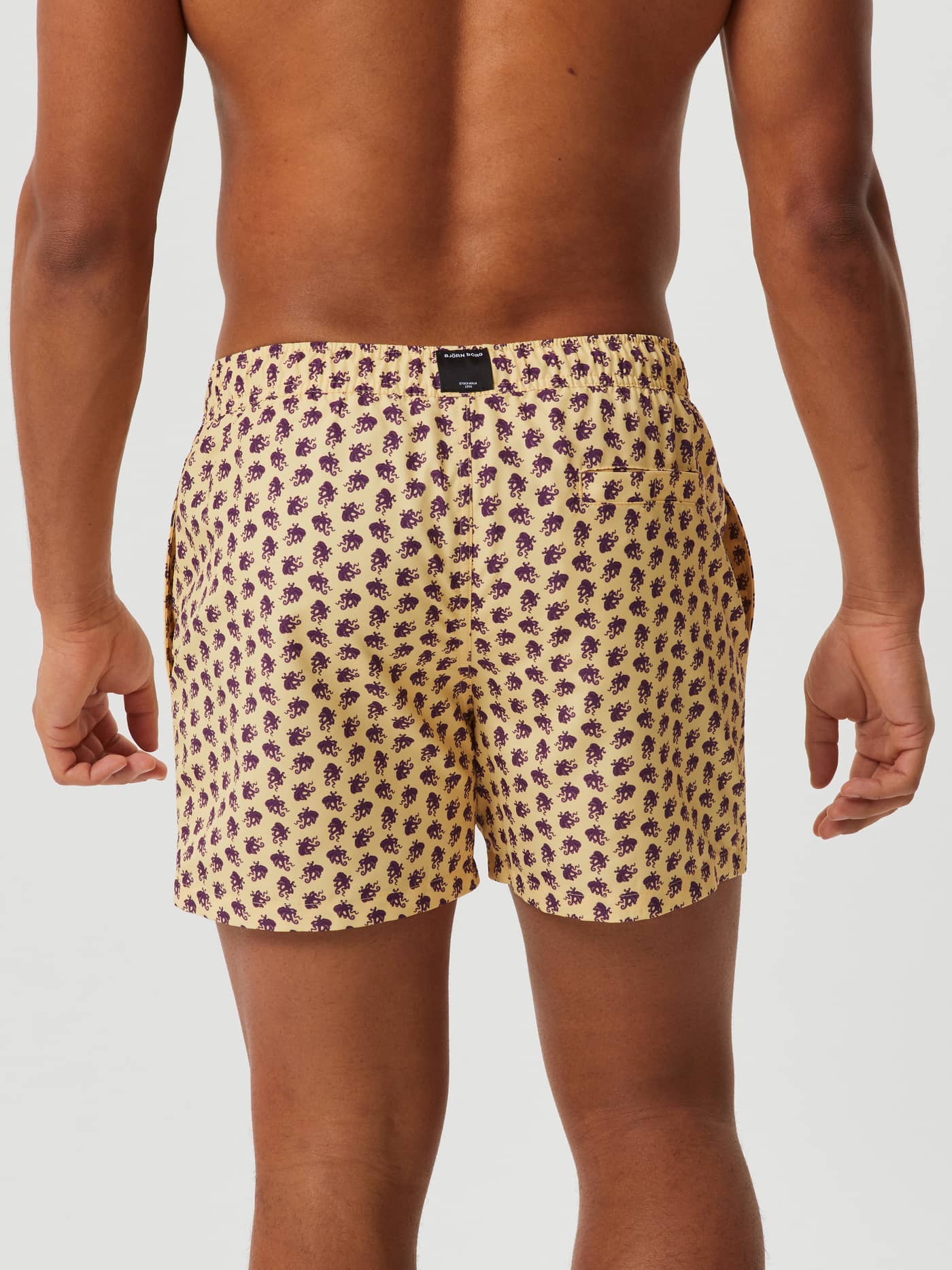 Back view of a man wearing the Björn Borg Print Swim Shorts in Octo 2 (Yellow) with a purple octopus print and black logo. - Image 3