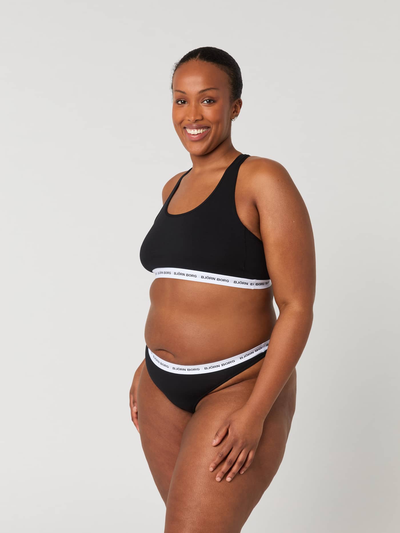 Side view of a model wearing the Core Logo Thong 2-pack in Black (Black) with a white waistband and black logo. - Image 3