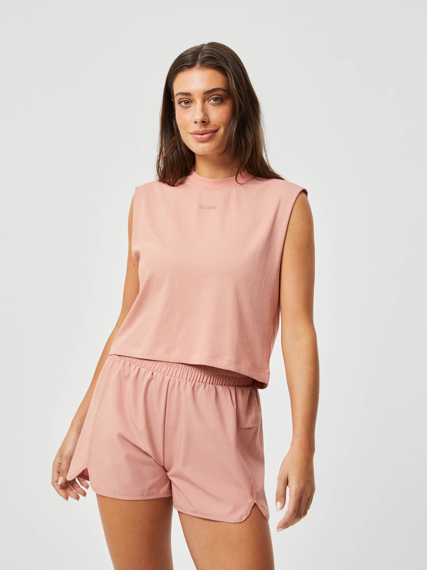 Front view of a woman in the loose-fit Borg Training Tank in Cameo Brown (Pink) with a matching logo for training. - Image 1