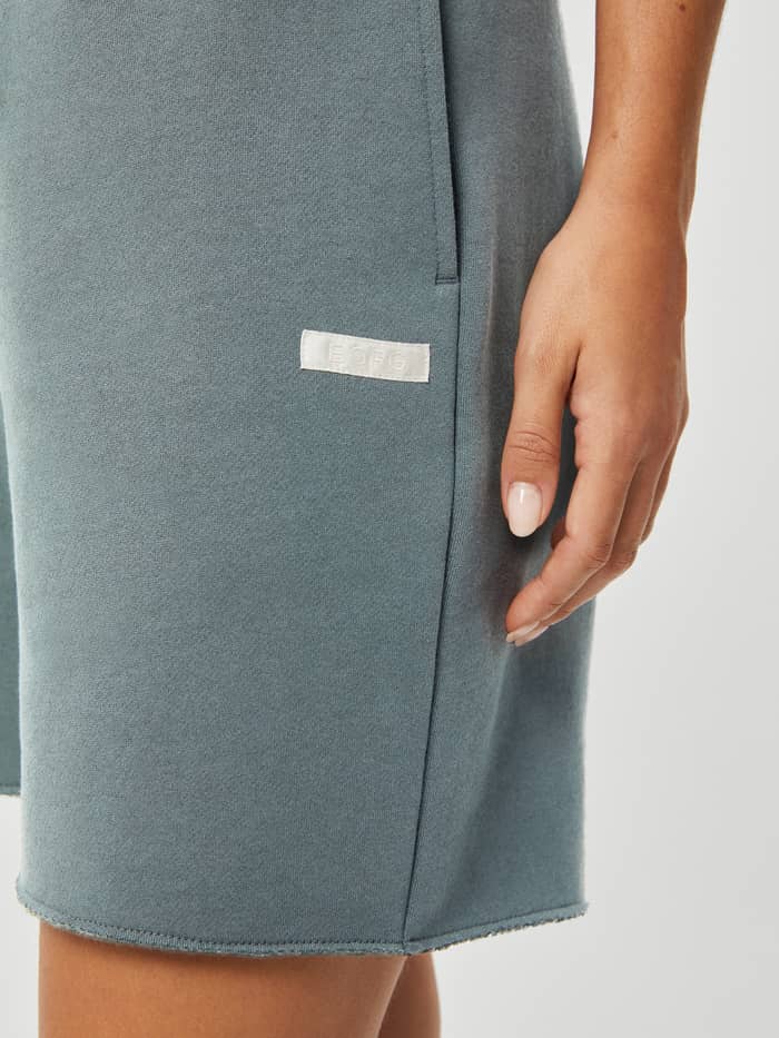 Close-up of the Studio Oversized Shorts in Gray Mist (Blue) showcasing the white satin BORG logo label and side pocket. - Image 4