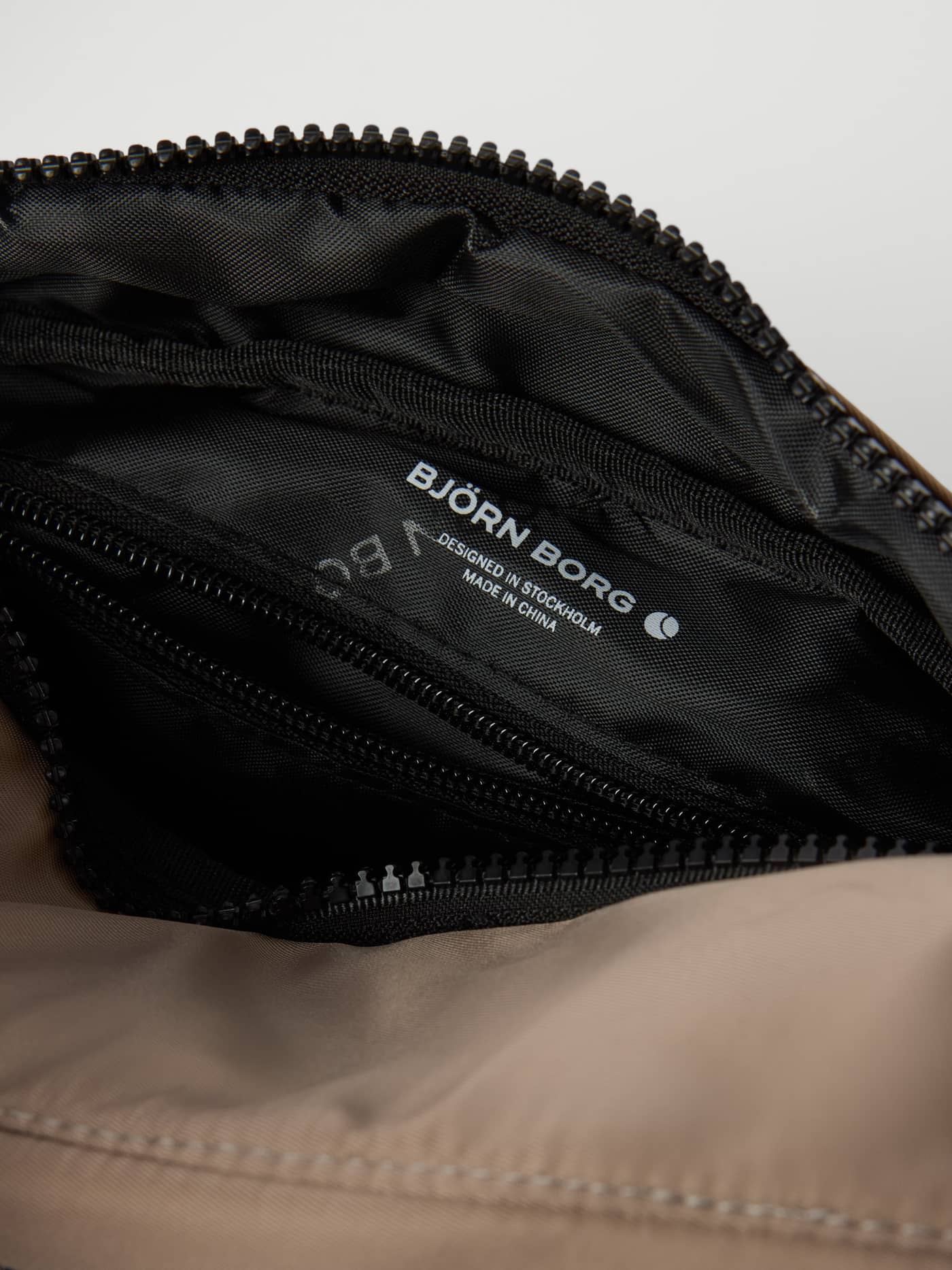 Detailansicht der innenliegenden Reißverschlusstasche und des weißen Logos der Borg Daily Crossover Bag in Fungi (Beige). - Image 3