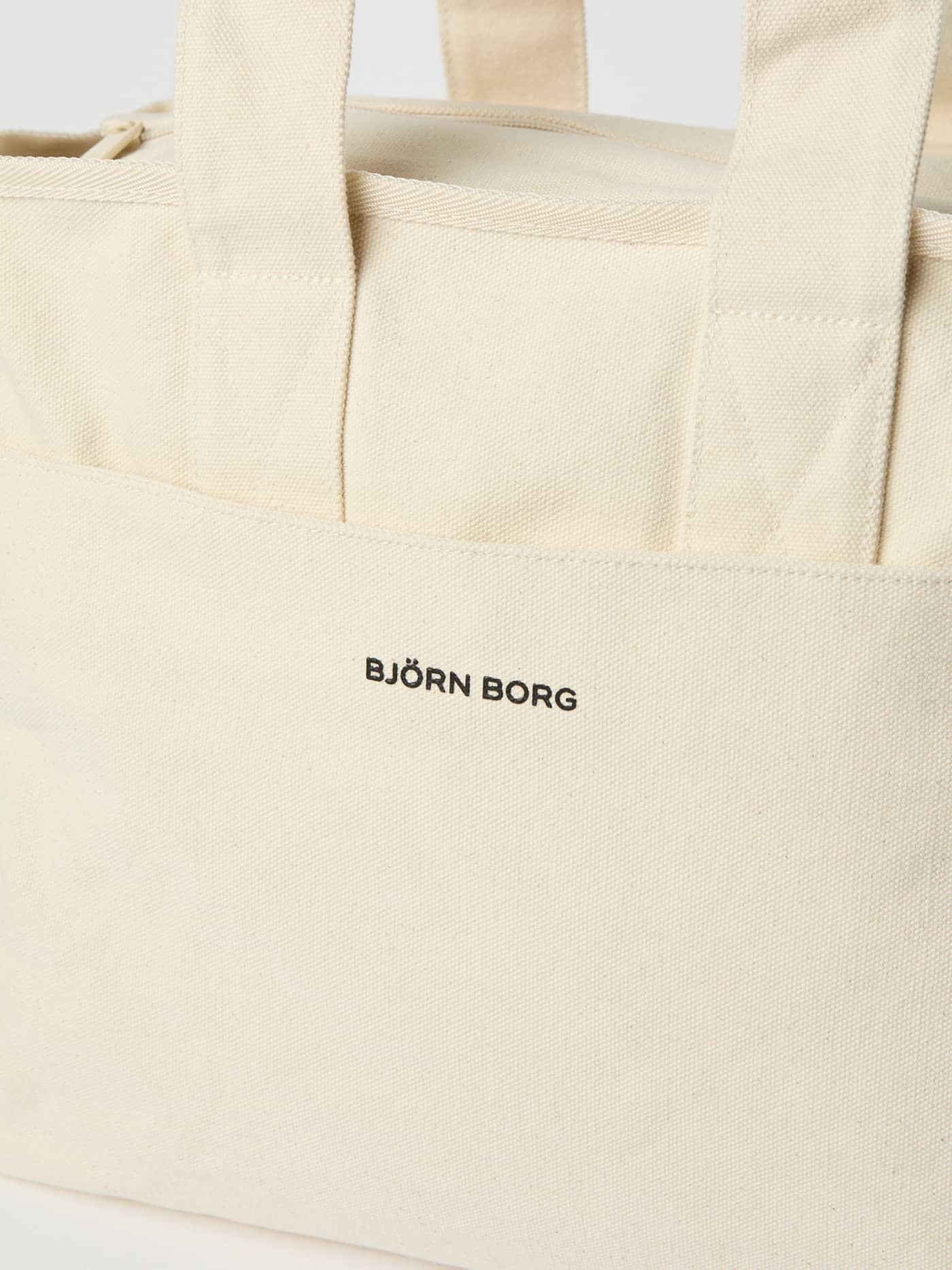 Close-up of the Studio Canvas Tote 27L in Egret (White) with a black logo on the durable cotton canvas front pocket. - Image 3