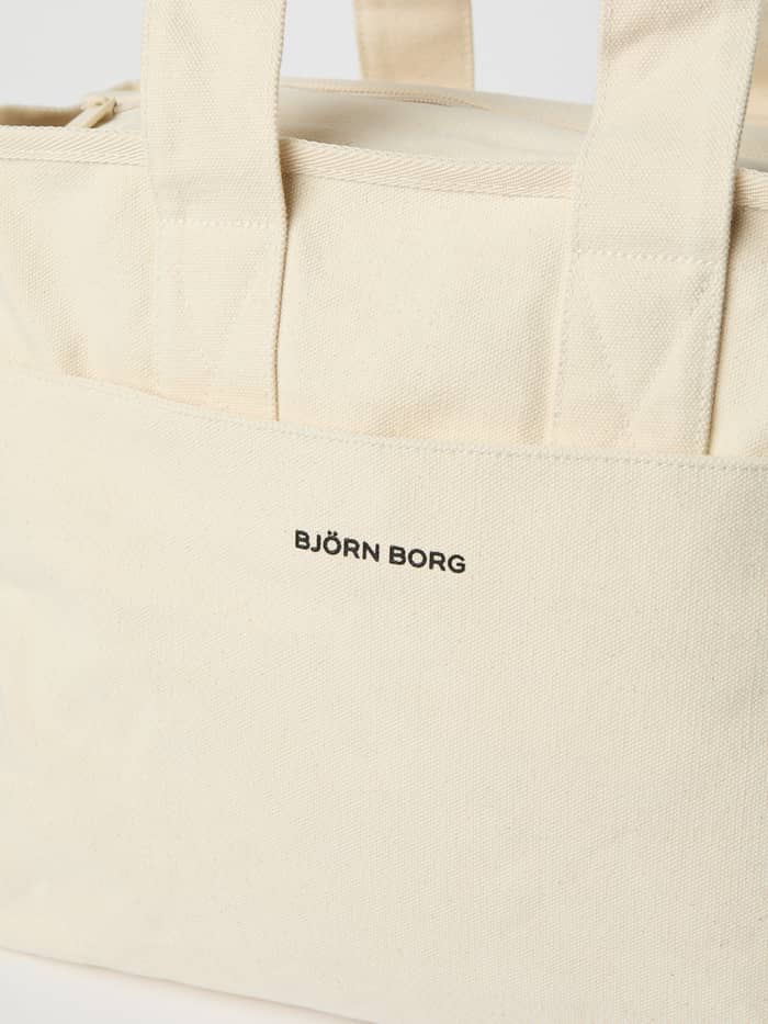 Close-up of the Studio Canvas Tote 27L in Egret (White) with a black logo on the durable cotton canvas front pocket. - Image 3