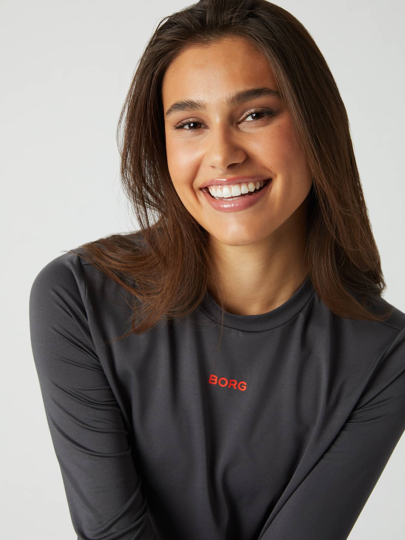 Close-up of a smiling woman wearing the asphalt grey Borg Long Sleeve T-shirt, showing the red logo on the chest. - Image 2