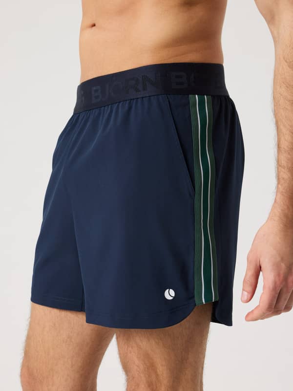 Side view of a man wearing the Ace Retro Shorts 6 Inch in Night sky (Navy) with retro stripes and a white tennis ball logo.