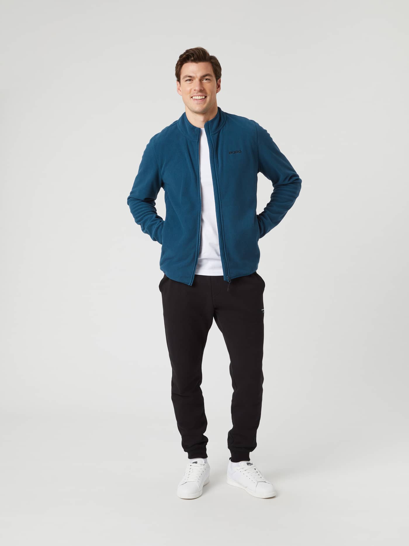 Full body front view of a man wearing the Björn Borg Borg Full Zip Fleece in blue, paired with black pants and white shoes. - Image 3