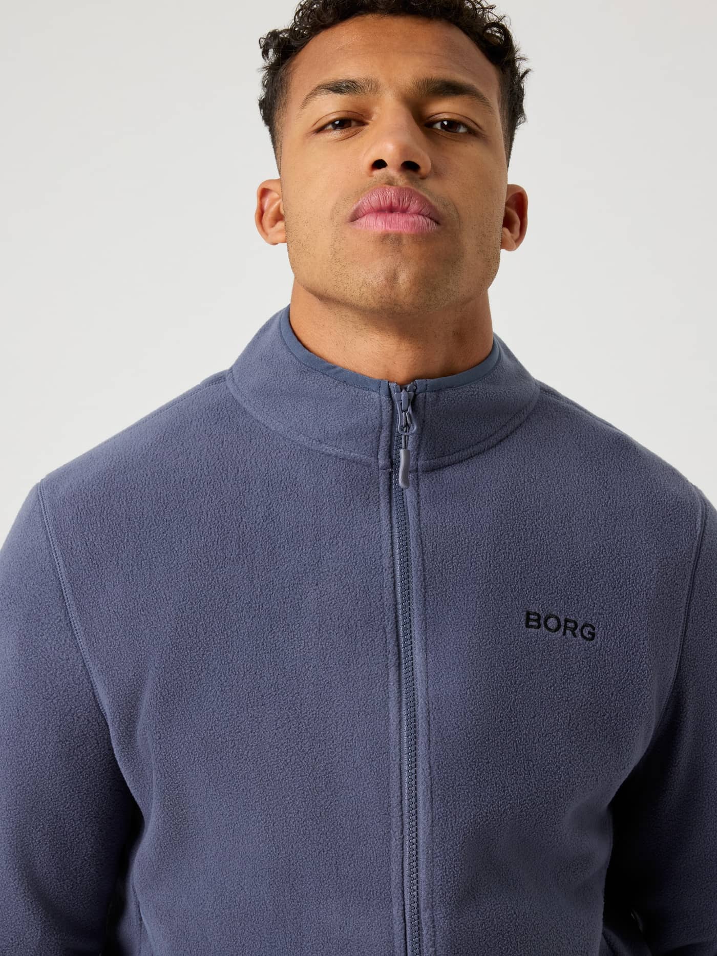 Close-up vooraanzicht van een model in de Borg Full Zip Fleece in Odyssey Gray (Blauw) met een zwart logo op de borst. - Image 4