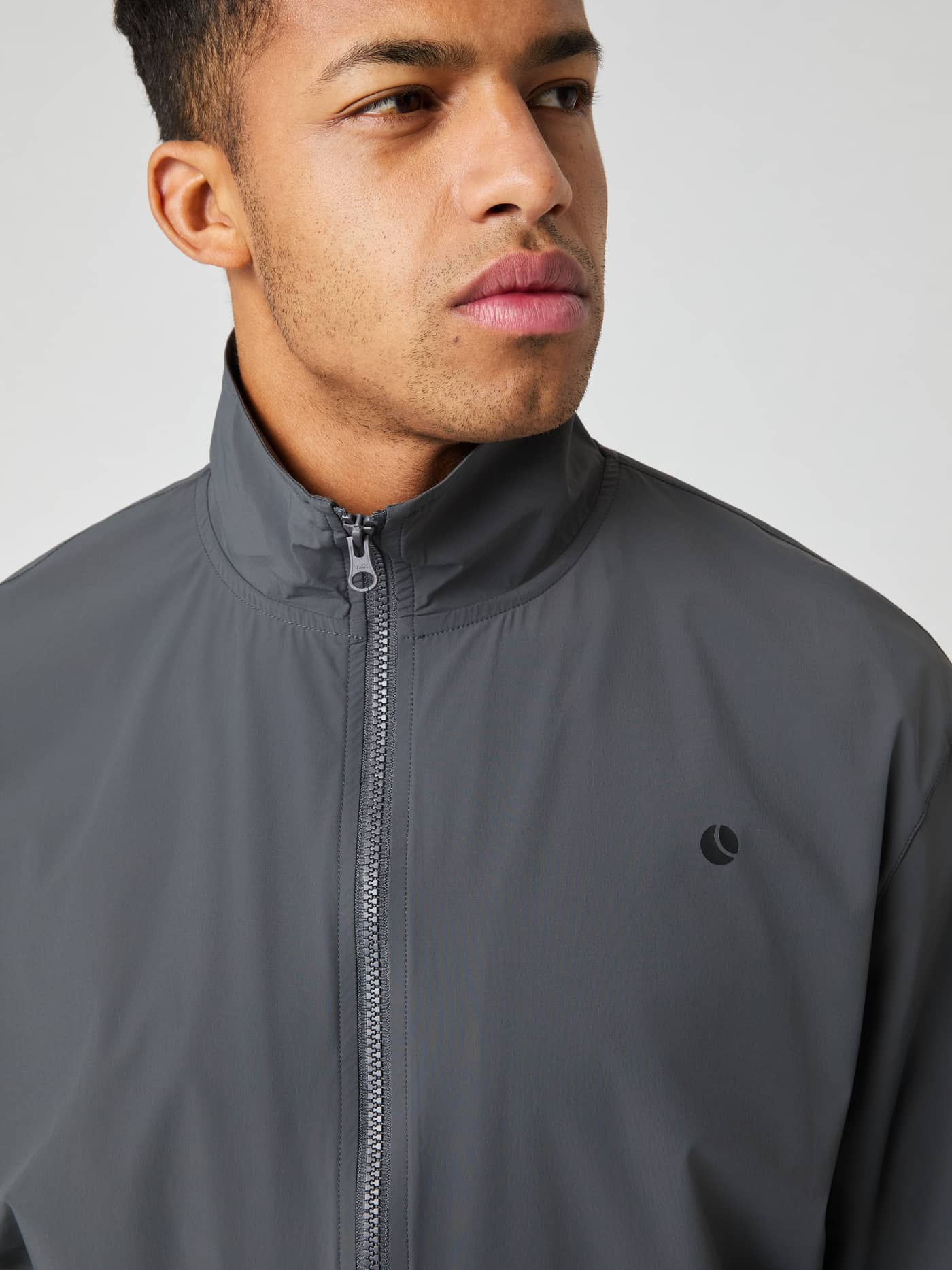 Close-up van een man in het functionele Ace Track Jacket in Asphalt (Grijs) met een kenmerkend tennisballogo. - Image 4