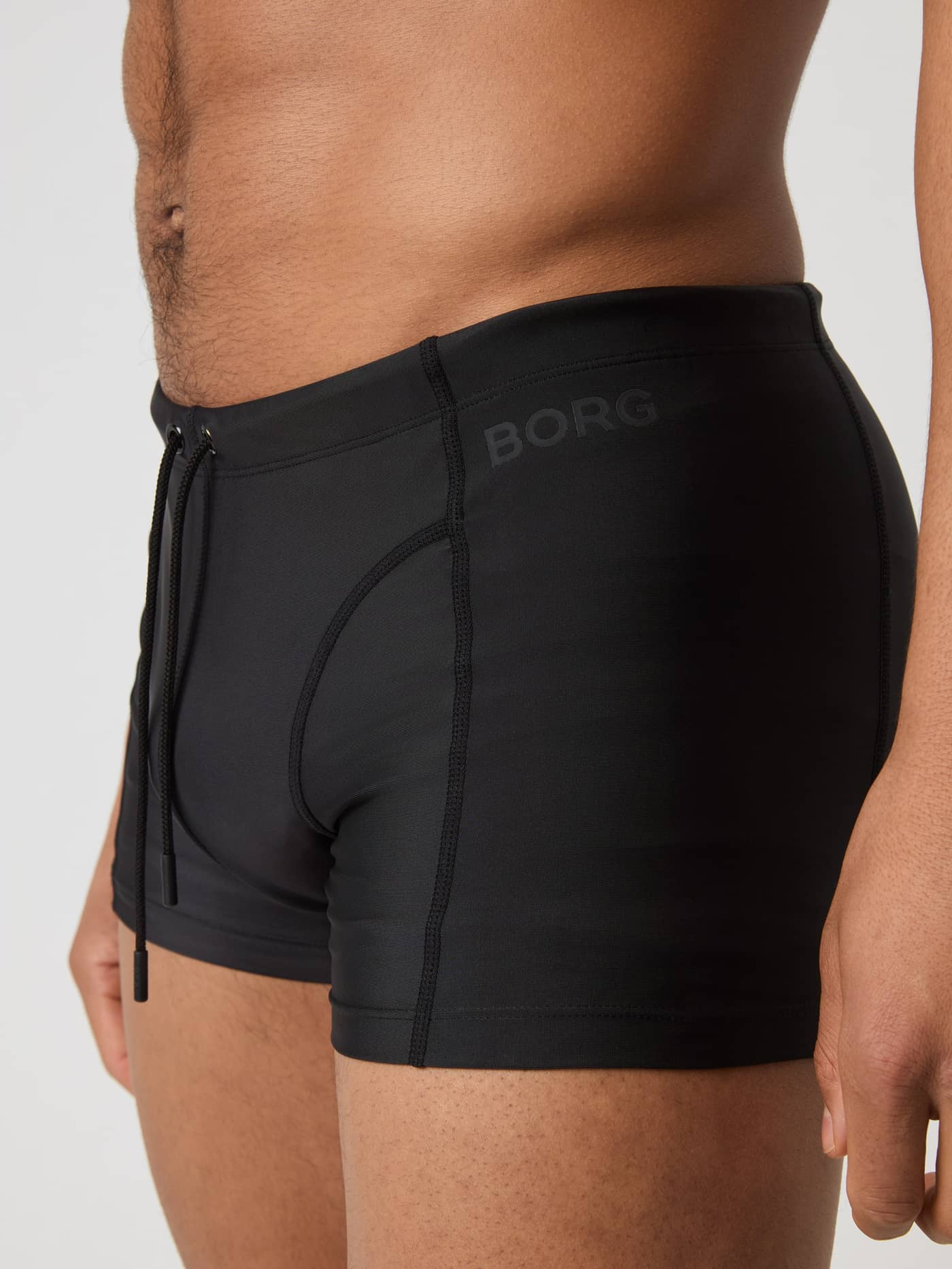 Side view of a man wearing the Borg Swim Trunks in Black Beauty (Black) featuring a slim fit and a black hip logo. - Image 4