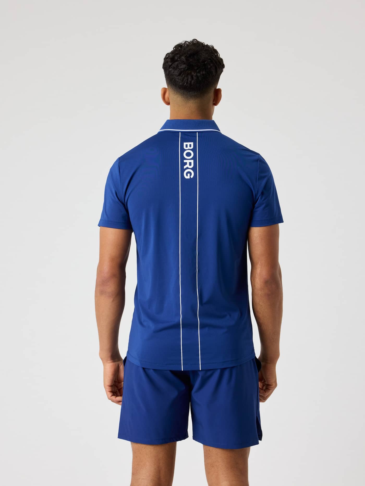 Ace Polo Shirt i Blue Depths (Blå) sedd bakifrån med en vit logorand för prestation på tennisbanan. - Image 2
