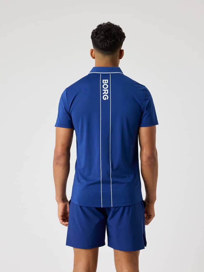 Back view of a man wearing the Ace Polo Shirt in Blue Depths (Blue) with a white logo stripe for performance tennis.