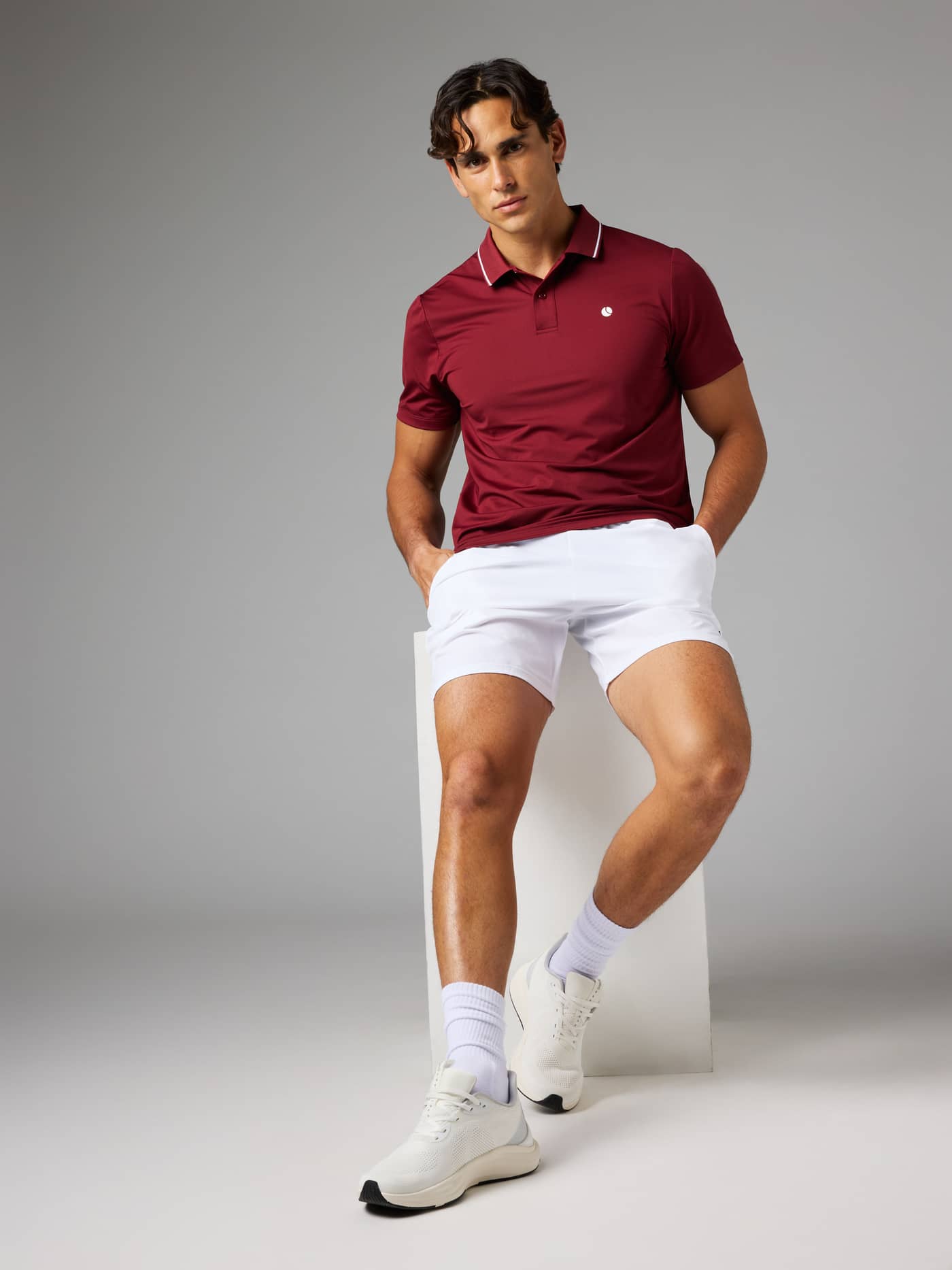Front view of a model sitting on a white cube, wearing the Björn Borg Ace Polo Shirt in Cabernet and white shorts for tennis. - Image 1