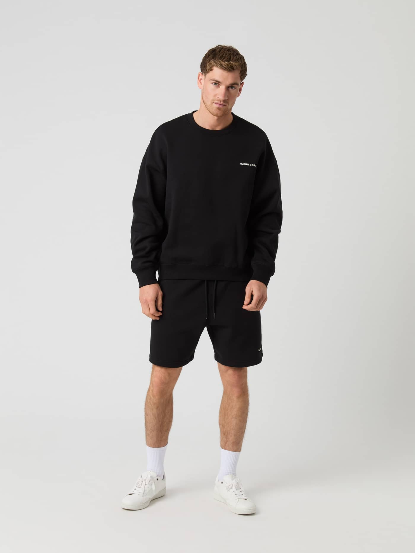 Full-body front view of a man wearing the Björn Borg Oversized Sweatshorts in Black Beauty (Black) with a white logo. - Image 3