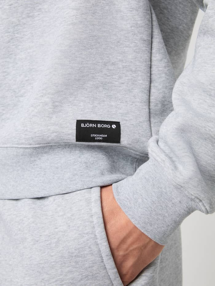 Detail van het Björn Borg Centre Sweatshirt in Light Grey Melange (Grijs) met een zwart geweven logolabel op de heup. - Image 5
