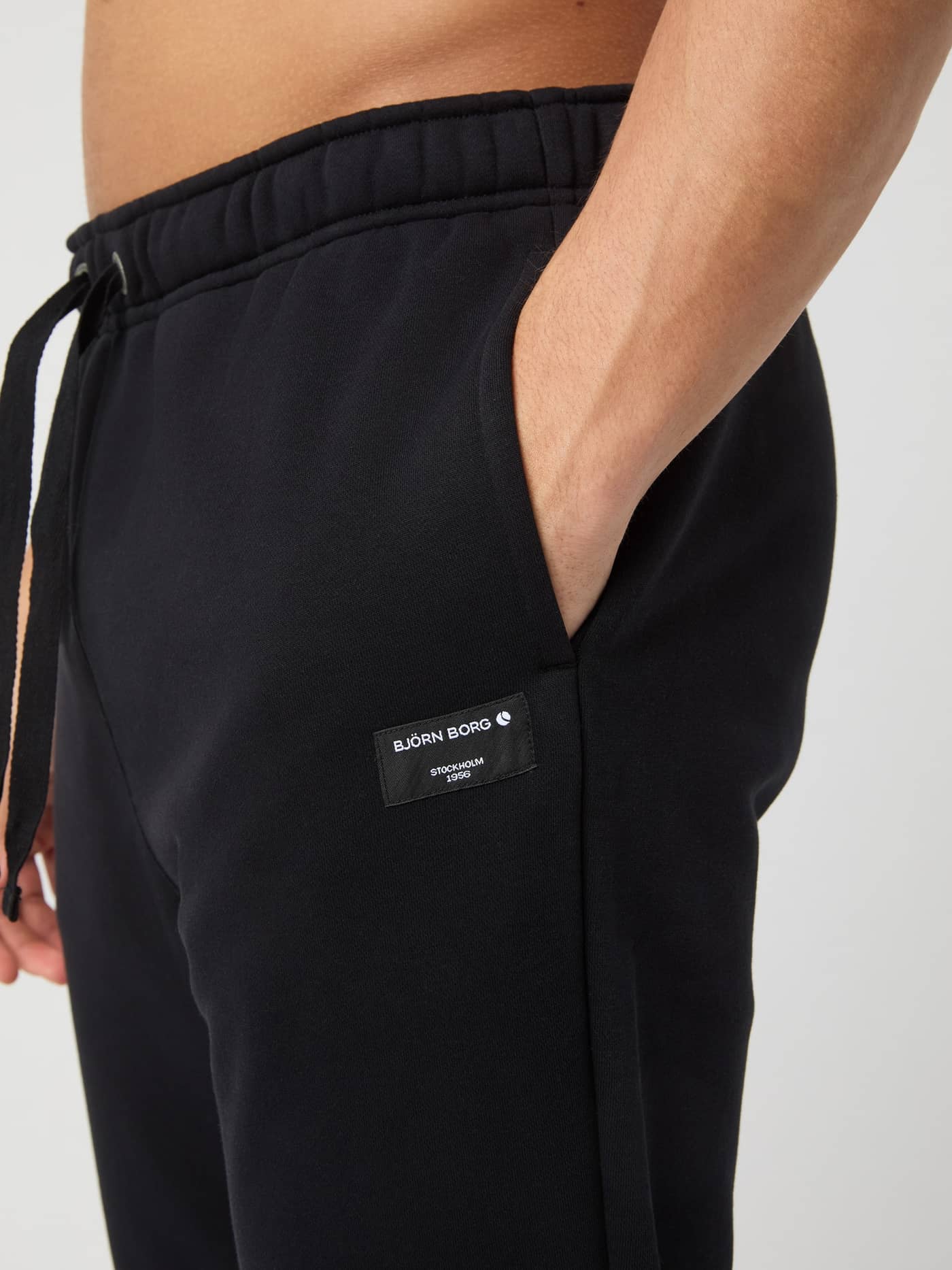 Centre Sweatpants i Black Beauty (Sort) i nærbillede med et hvidt vævet logomærke og hånden i lommen. - Image 4
