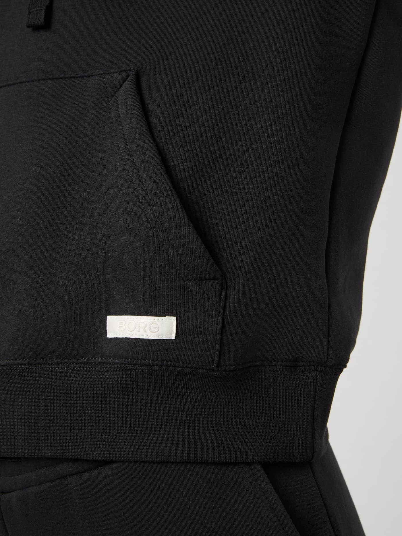 Détail de la poche kangourou et du logo tissé blanc du Centre Hoodie surdimensionné en Black Beauty (Noir). - Image 5