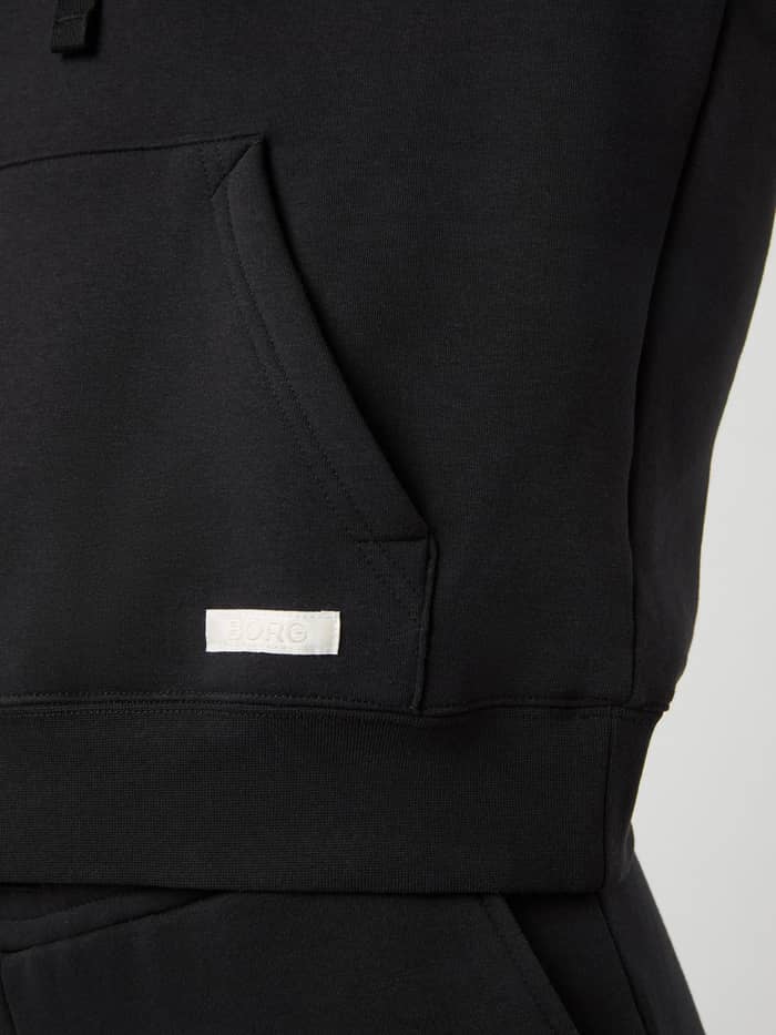 Detalje af kængurulomme og hvidt vævet logo på Centre Hoodie til kvinder i Black Beauty (Sort) med oversized pasform. - Image 5