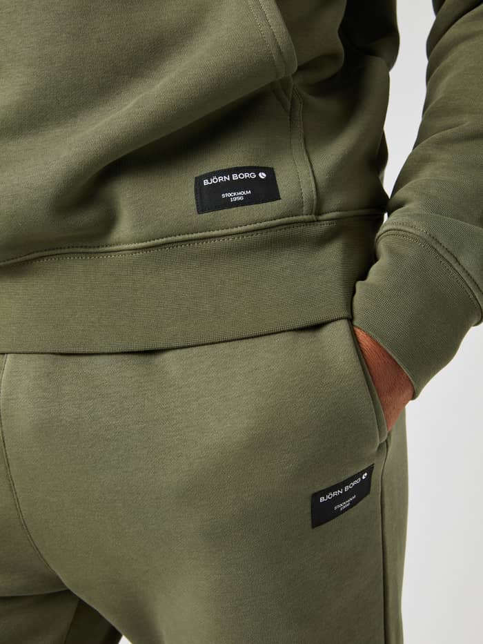 Close-up of the Björn Borg Centre Hoodie in dusty olive, showing the woven logo label above the ribbed hem. - Image 4