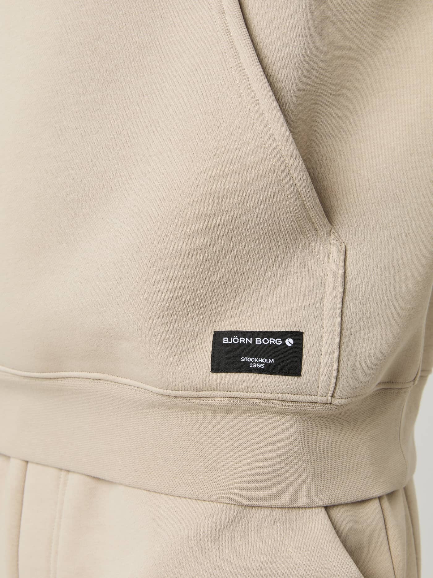 Close-up of the beige Björn Borg Centre Hoodie, focusing on the kangaroo pocket and black woven logo label at the hem. - Image 5