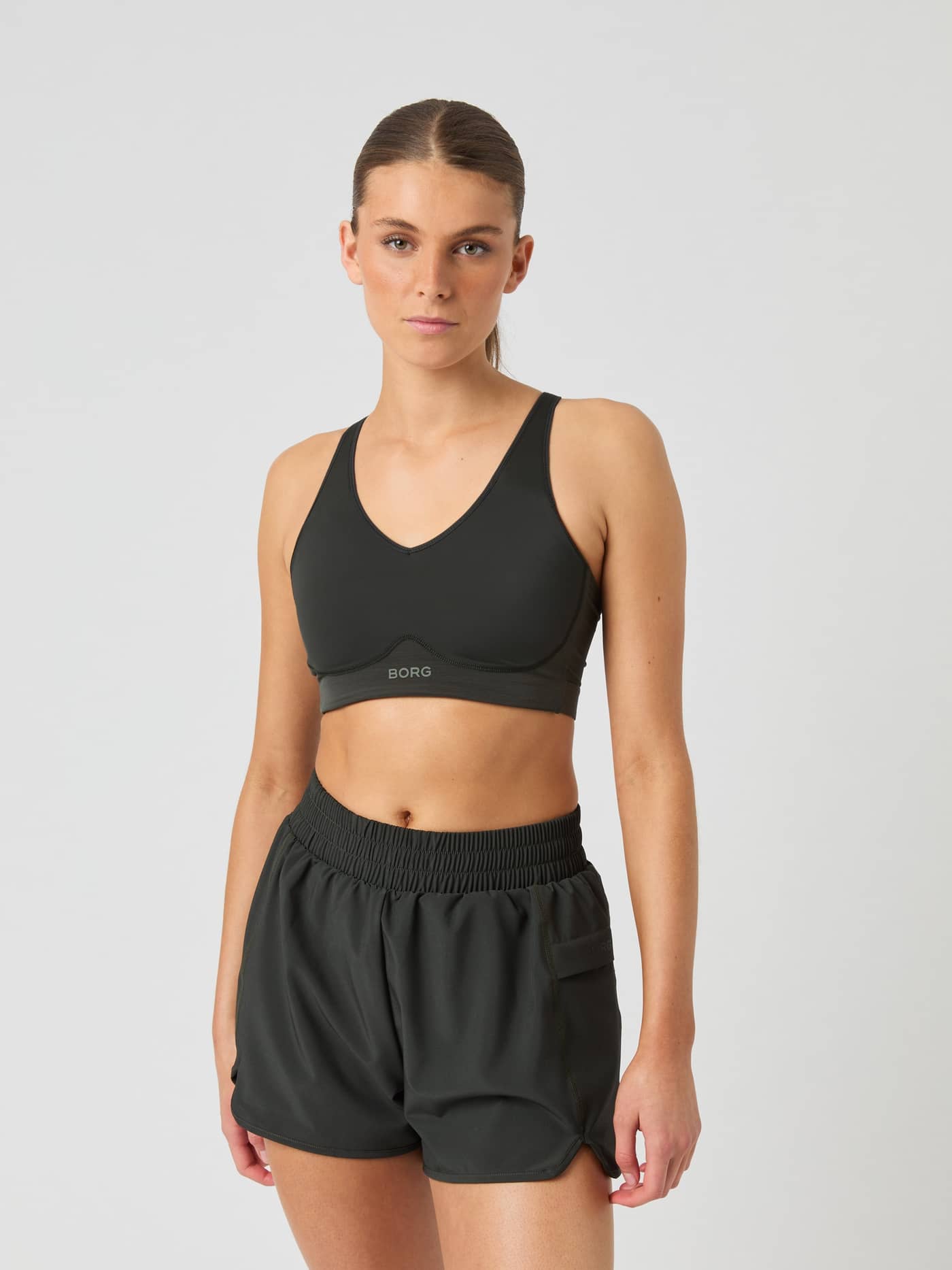 Front view of a model in the green Borg Mid V Support Sports Bra for training, featuring a grey logo and a supportive fit. - Image 3