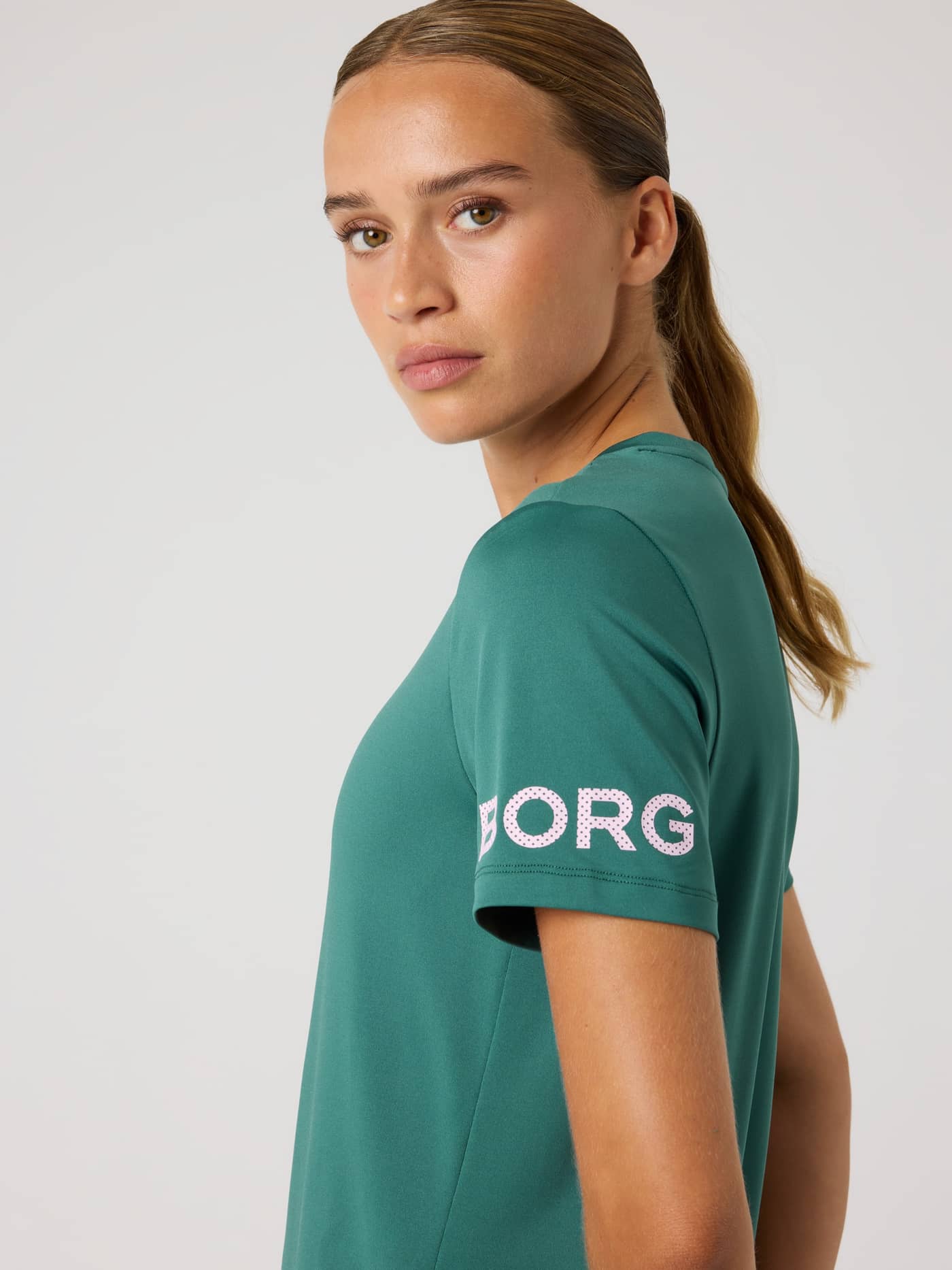 Seitenansicht eines Models im Mallard Green (Grün) Borg T-Shirt fürs Training mit pinkem Logo am Ärmel und stilvollem Fit. - Image 1