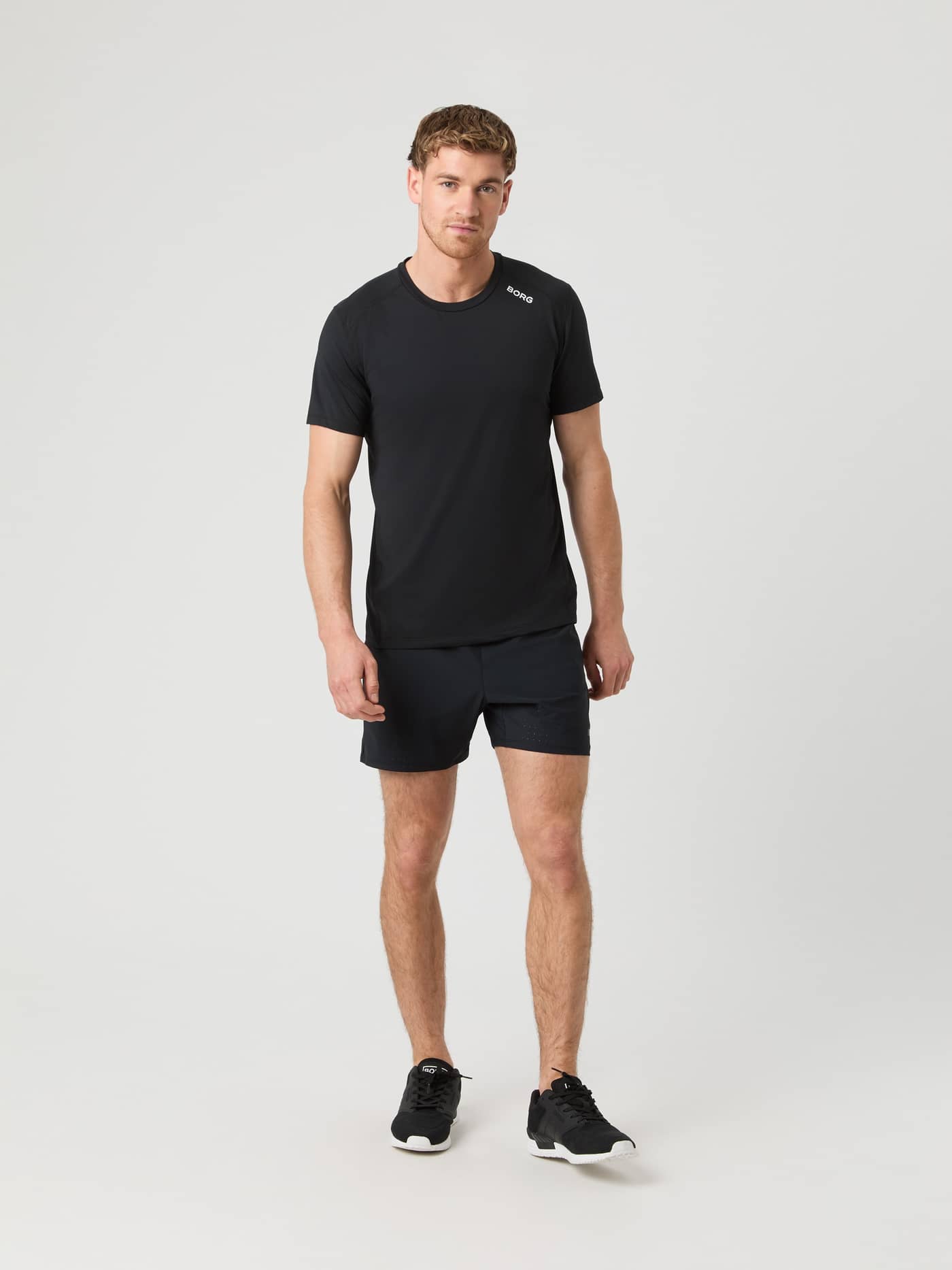 Male model standing in a studio wearing the black Borg Athletic T-shirt with matching black shorts and sneakers. - Image 3