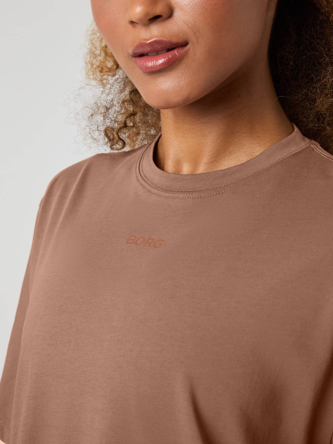 Gros plan du Studio Oversized T-Shirt pour femme en Acorn (Marron) montrant le col rond et le petit logo marron. - Image 4