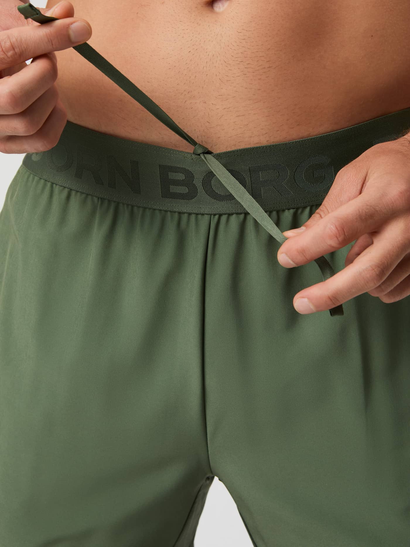 Close-up of a man tying the drawstring on the thyme green Björn Borg shorts, highlighting the logo waistband. - Image 6