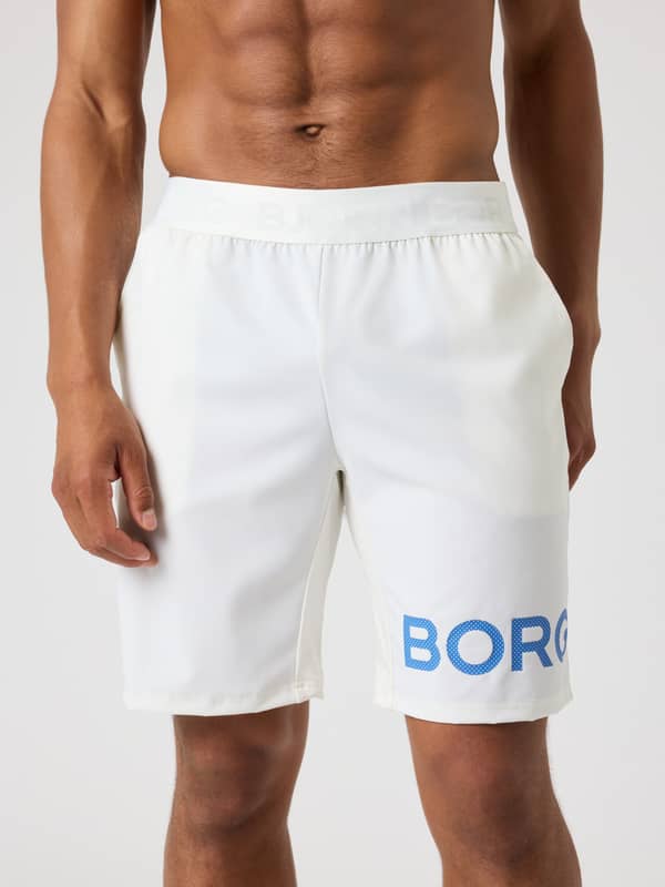 Vue de face d'un homme portant le Borg Shorts 9 Inch en Tofu (Blanc) avec un logo bleu perforé pour l'entraînement.