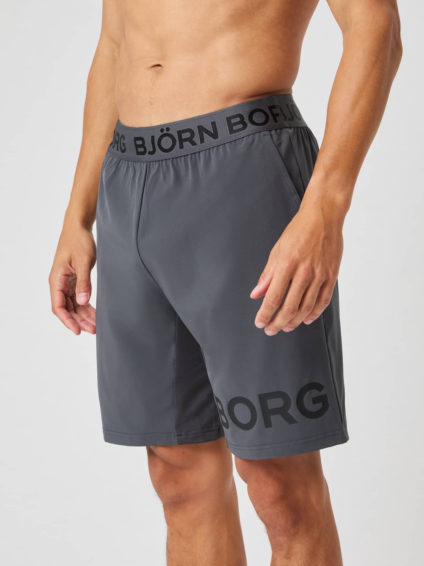 Side-front view of a model in dark grey Björn Borg 9" Shorts, showing the logo waistband and BORG print on the leg. - Image 1