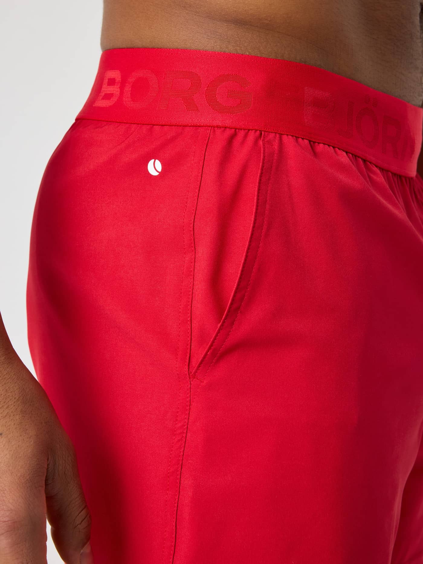 Close-up of the men's Borg Shorts 6 Inch in Lychee (Red), training shorts with a red logo waistband and white tennis icon. - Image 4