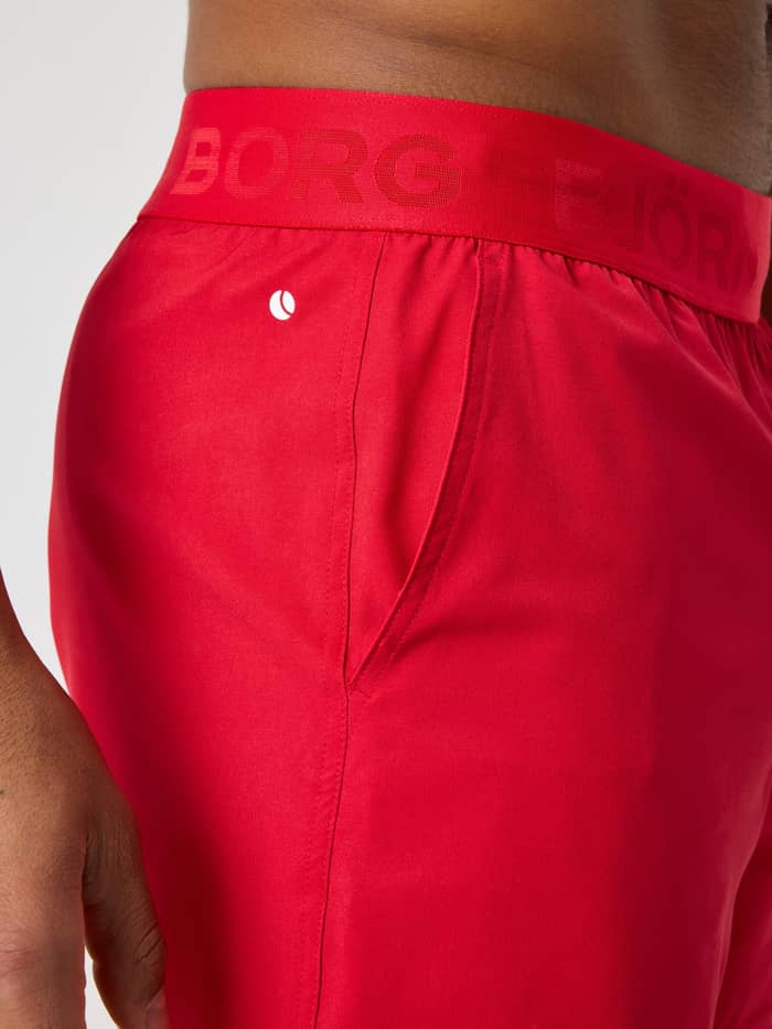 Close-up of the men's Borg Shorts 6 Inch in Lychee (Red), training shorts with a red logo waistband and white tennis icon. - Image 4