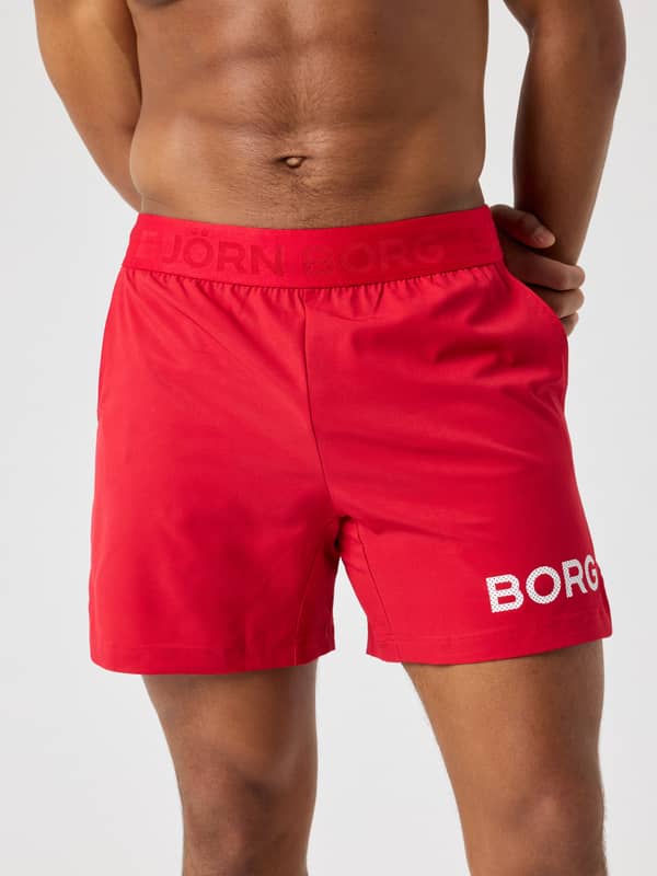Front view of a man wearing the Borg Shorts 6 Inch in Lychee (Red) with a white logo for tennis and training performance.
