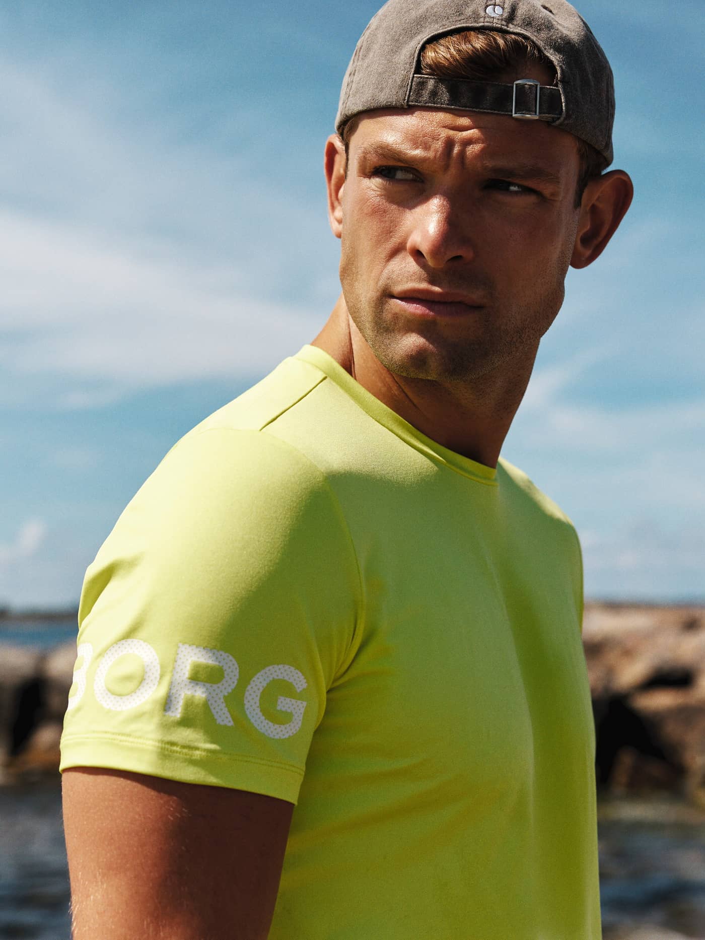 Close-up of a man wearing the bright yellow Borg T-Shirt and a cap, looking over his shoulder outdoors. - Image 1