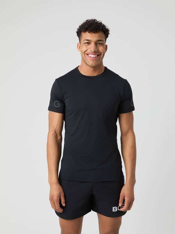 Ein lächelnder Mann im Borg T-Shirt in Black Tonal Logo (Schwarz) für das Training mit 4-Wege-Stretch und tonalem Logo.